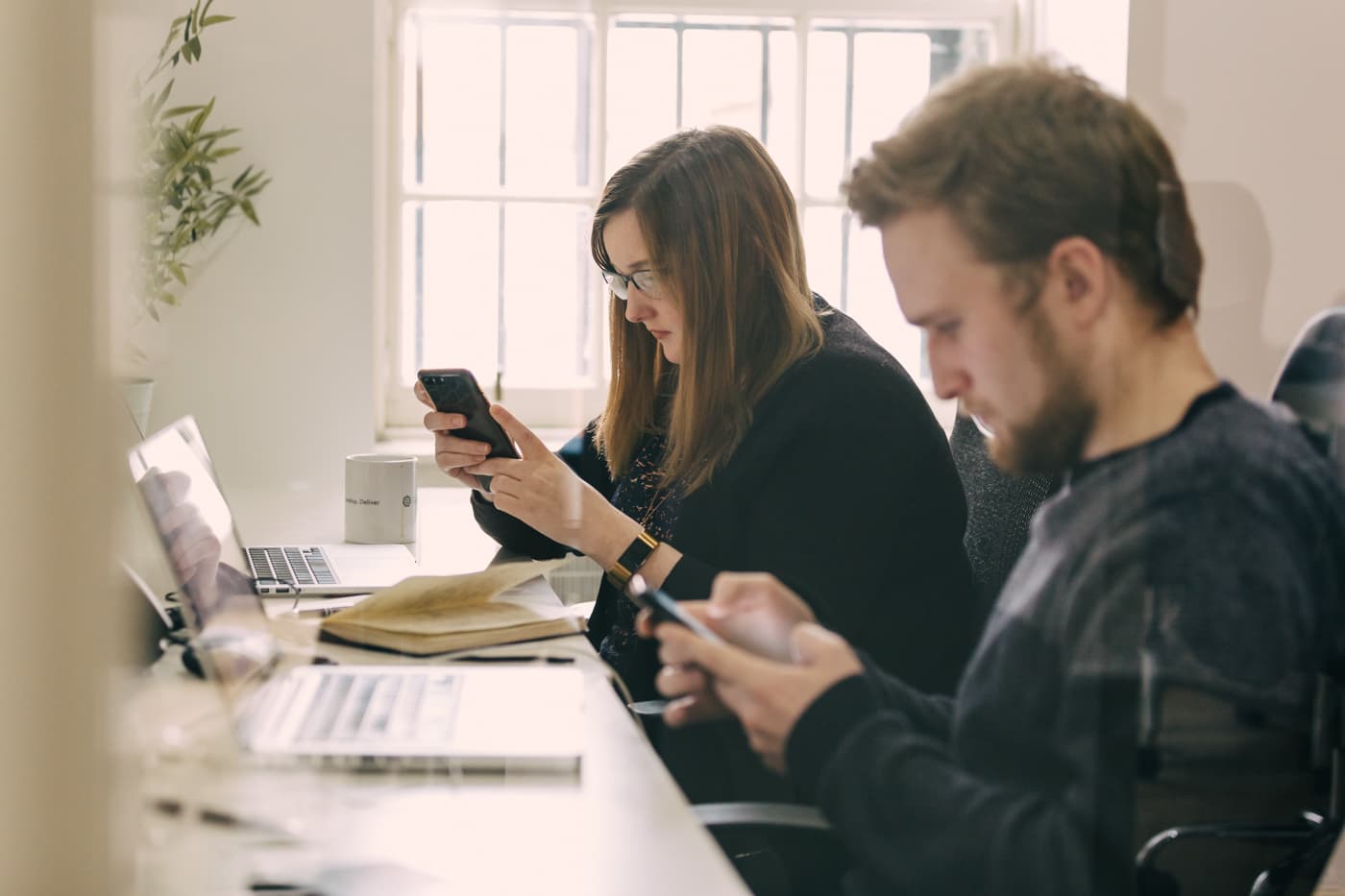 Two people working side by side at a desk, one reviewing a product on a phone and one taking notes